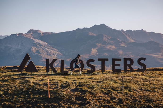 A trail runner runs in front of the monastery logo on the Gotschna