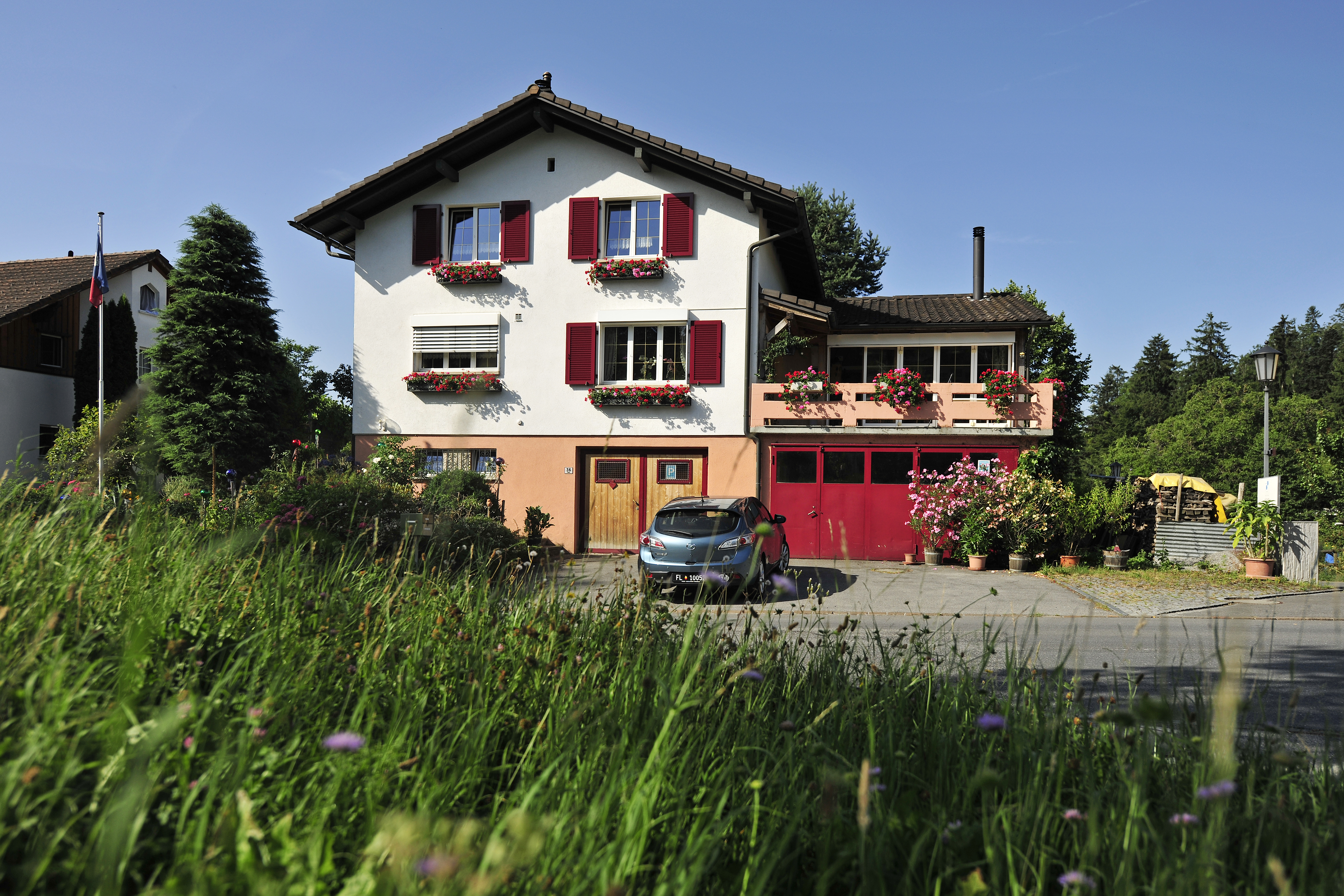 Charmante Außenansicht des Bed & Breakfast Büchel mit roten Fensterläden und Blumenwiese im Vordergrund.